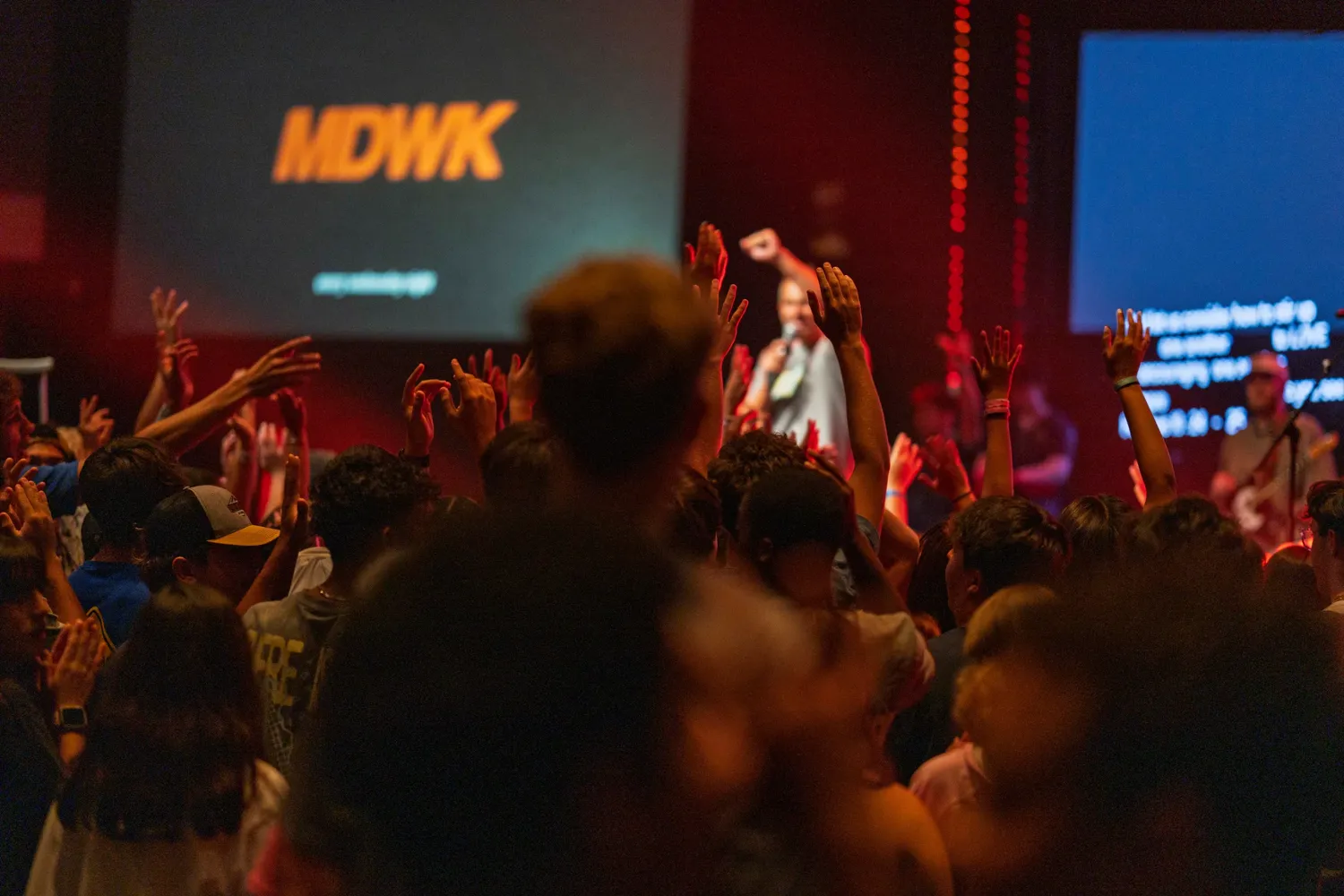 Students worshipping with hands raised at Student MDWK, with MDWK on the screen behind them